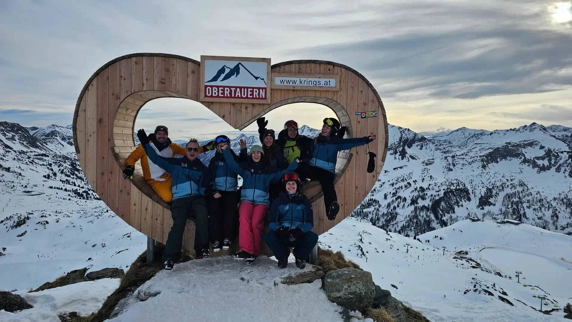 Eine Gruppe von acht Personen in Winterkleidung posiert zusammen in einem großen hölzernen Herzrahmen mit der Aufschrift "Obertauern", umgeben von verschneiten Bergen und bewölktem Himmel in einer malerischen Alpenlandschaft.