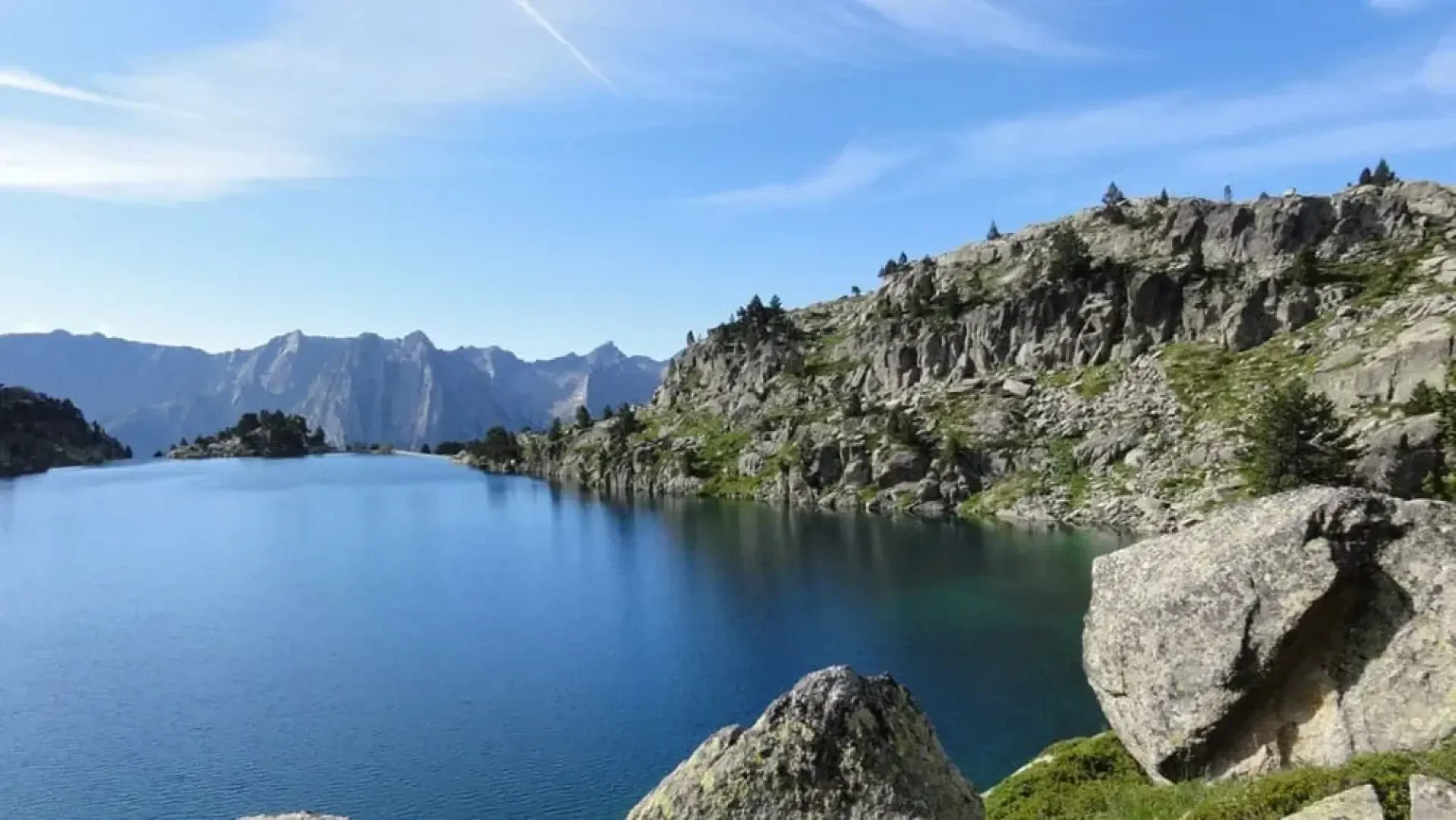 Ein klarer, blauer Bergsee, gesäumt von felsigen Klippen und vereinzelten Pinienbäumen unter einem hellen, teilweise bewölkten Himmel mit fernen, zerklüfteten Gipfeln im Hintergrund.