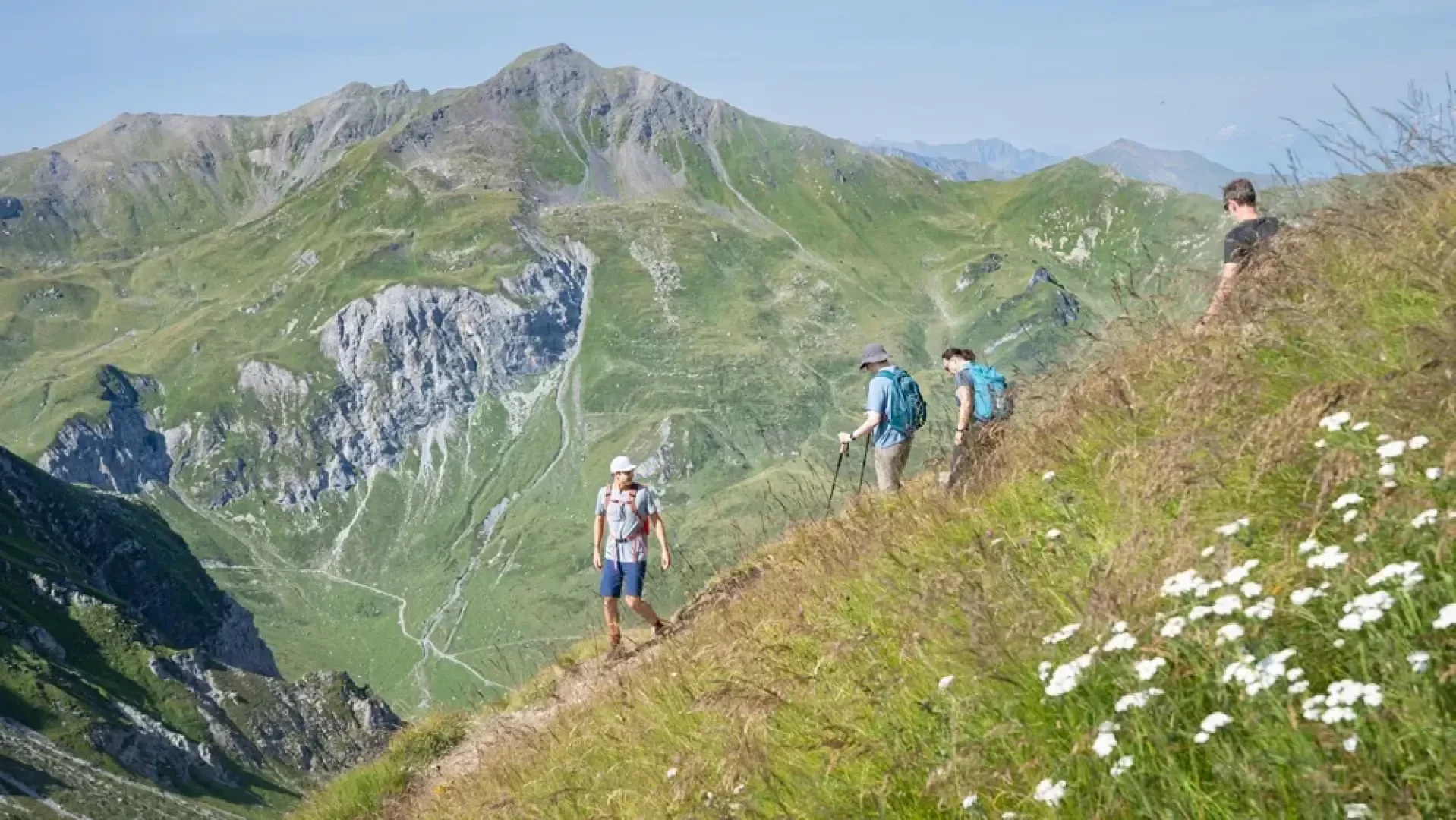 Vier Wanderer gehen auf einem grasbewachsenen Bergpfad mit Wildblumen, hoch über einem grünen Tal, umgeben von schroffen, steilen Hängen und fernen Gipfeln unter einem klaren blauen Himmel.