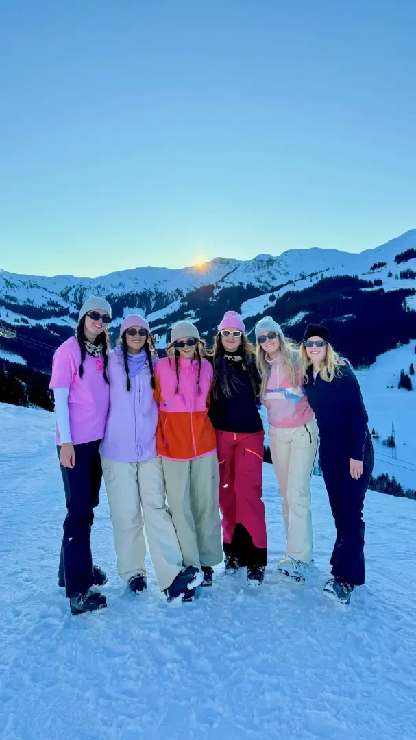 Sechs Frauen in bunter Winterkleidung stehen Arm in Arm auf einem verschneiten Berg mit einem Sonnenuntergang und schneebedeckten Gipfeln im Hintergrund. Alle lächeln, tragen Sonnenbrillen und sind warm gekleidet.