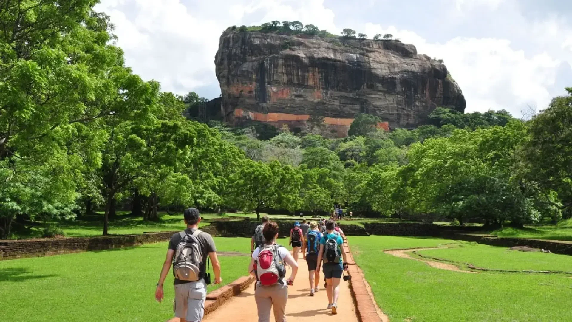 Eine Gruppe von Touristen geht bei teilweise bewölktem Himmel einen von üppigem Grün umgebenen Weg in Richtung des Sigiriya-Felsens, einer großen alten Felsenfestung in Sri Lanka, entlang.