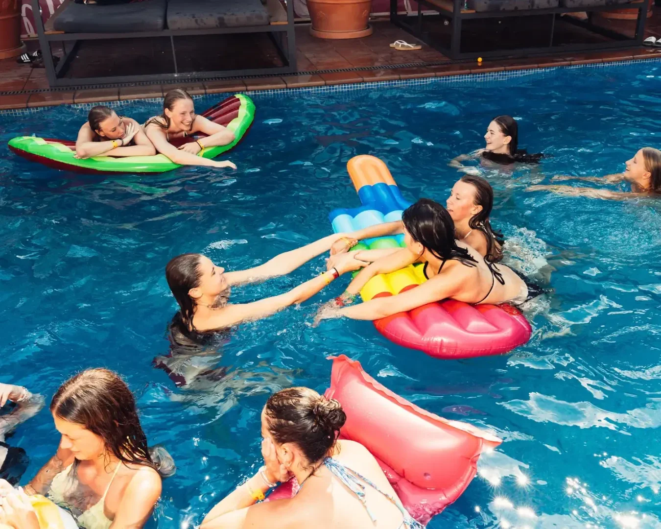 Eine Gruppe von Frauen in Badeanzügen genießt einen sonnigen Tag in einem Schwimmbad, lächelt, plaudert und spielt auf bunten aufblasbaren Flößen und Schwimmflügeln.