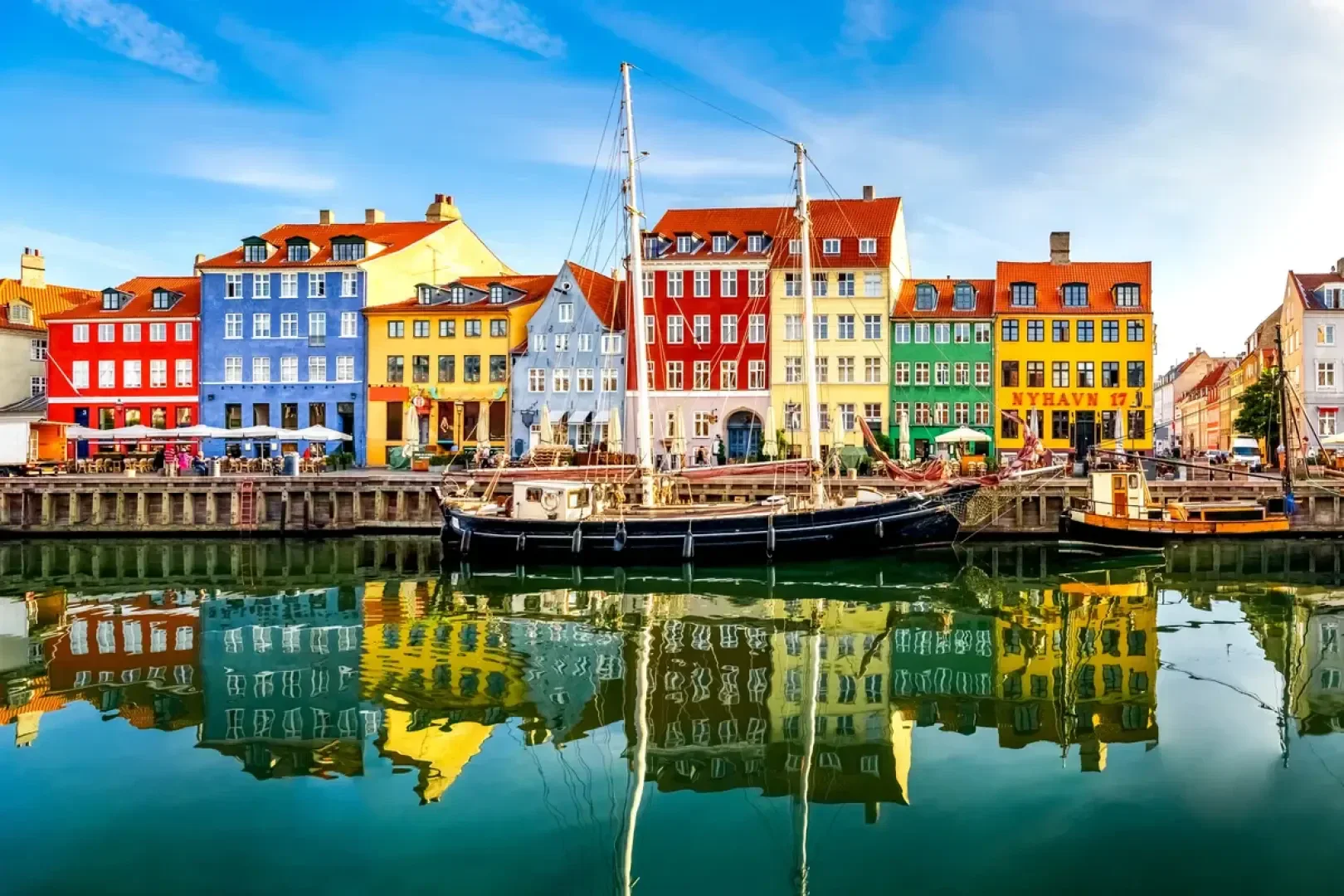 Bunte Gebäude säumen die Uferpromenade Nyhavn in Kopenhagen, Dänemark. Entlang des Kanals liegen Boote vor Anker, die sich im ruhigen Wasser unter blauem Himmel spiegeln.