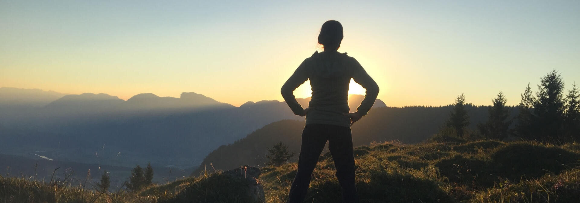 Eine Person steht auf einem grasbewachsenen Gelände, die Hände in die Hüften gestemmt, mit Blick auf den Sonnenaufgang oder -untergang über fernen Bergen und Tälern, die sich als Silhouette gegen den Himmel abheben.