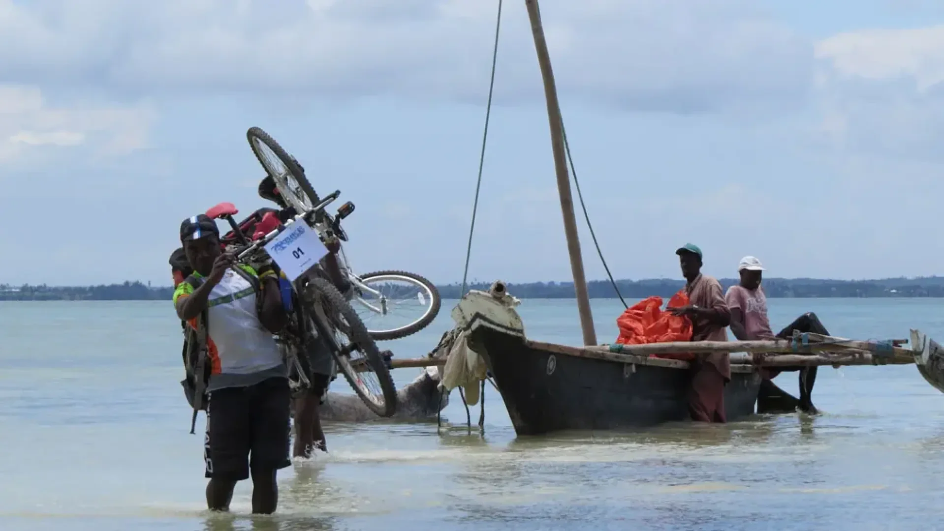 Ein Mann watet mit einem Fahrrad durch seichtes Wasser, während drei Personen in einem Holzboot in der Nähe unter einem teilweise bewölkten Himmel sitzen.