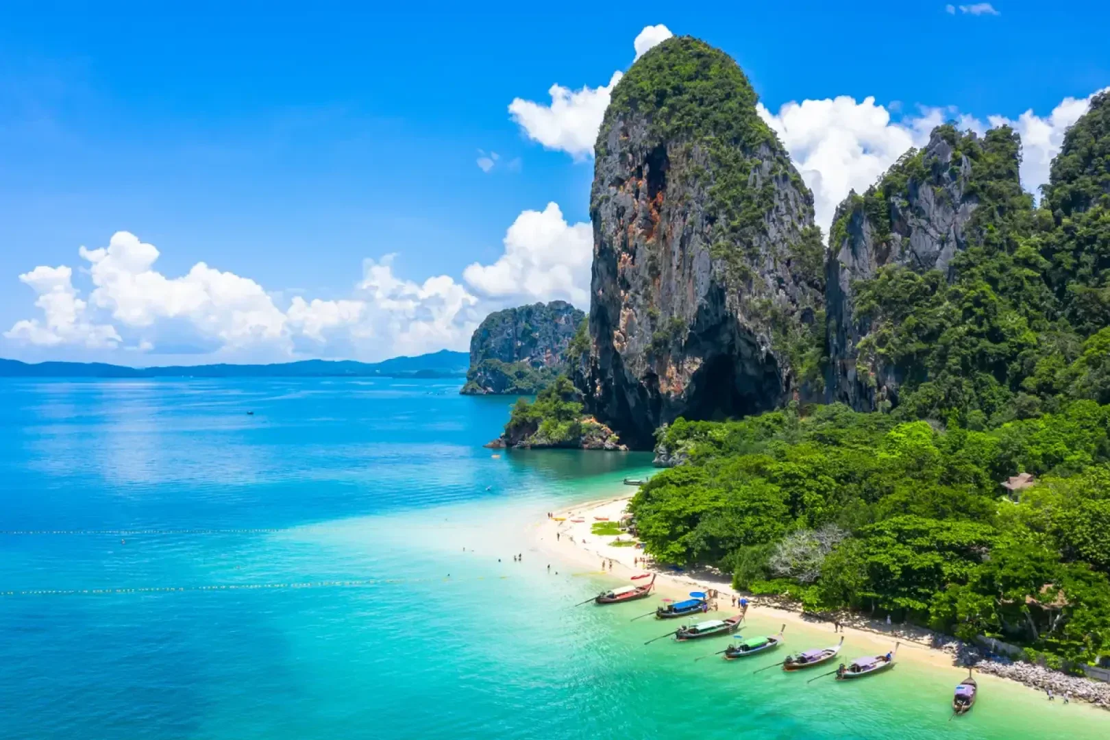Ein tropischer Strand mit türkisfarbenem Wasser, weißem Sand, traditionellen Booten, üppig grünen Bäumen und einer dramatischen Kalksteinklippe unter einem strahlend blauen Himmel mit vereinzelten Wolken.