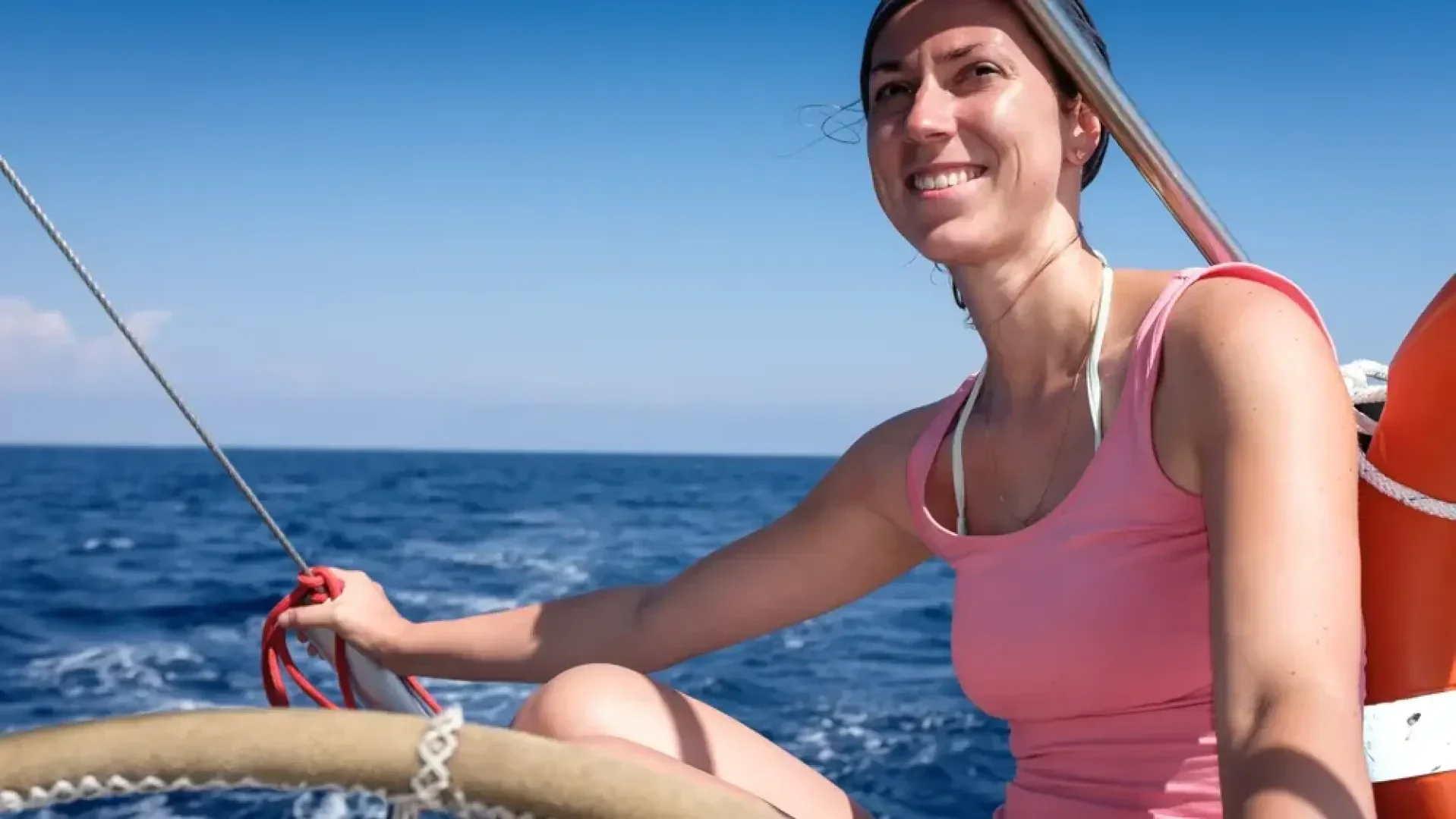 Eine Frau, die ein rosa Tank-Top und ein Stirnband trägt, lächelt beim Segeln auf einem Boot und hält ein Seil. Der blaue Ozean und der klare Himmel sind im Hintergrund zu sehen.