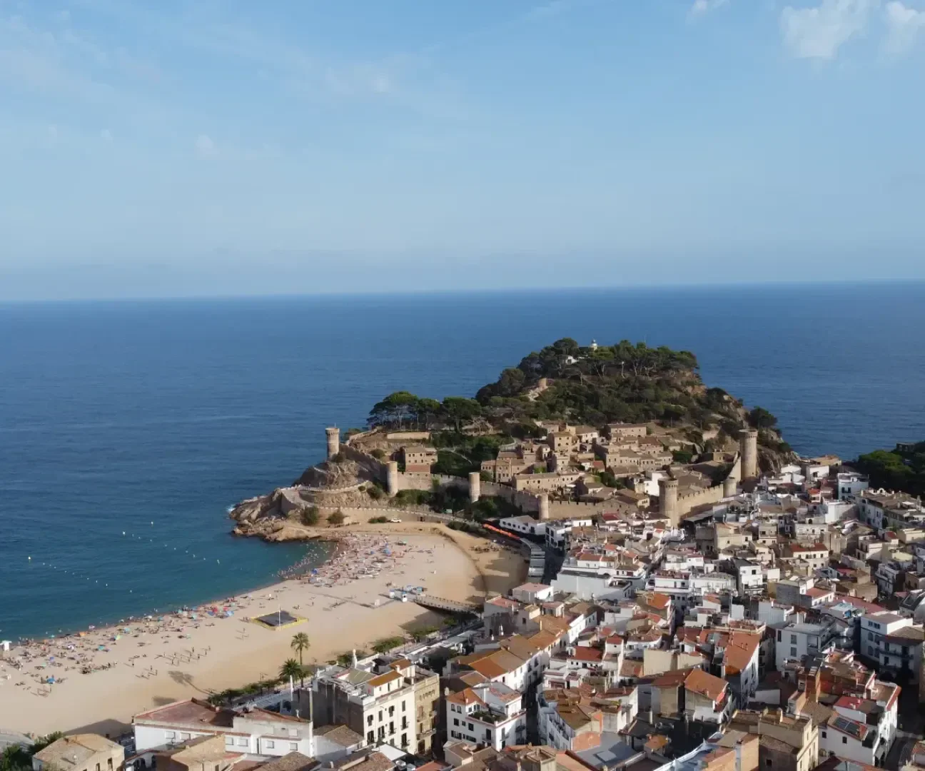 Luftaufnahme einer Küstenstadt mit roten Dächern, einem Sandstrand und einer historischen Festung auf einem Hügel mit Blick auf das blaue Meer unter einem klaren Himmel.