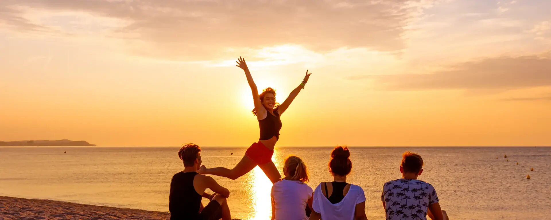 Fünf Personen sitzen bei Sonnenuntergang im Sand am Meer. Eine Person springt freudig mit erhobenen Armen vor dem goldenen Himmel und dem ruhigen Wasser vor die Gruppe.