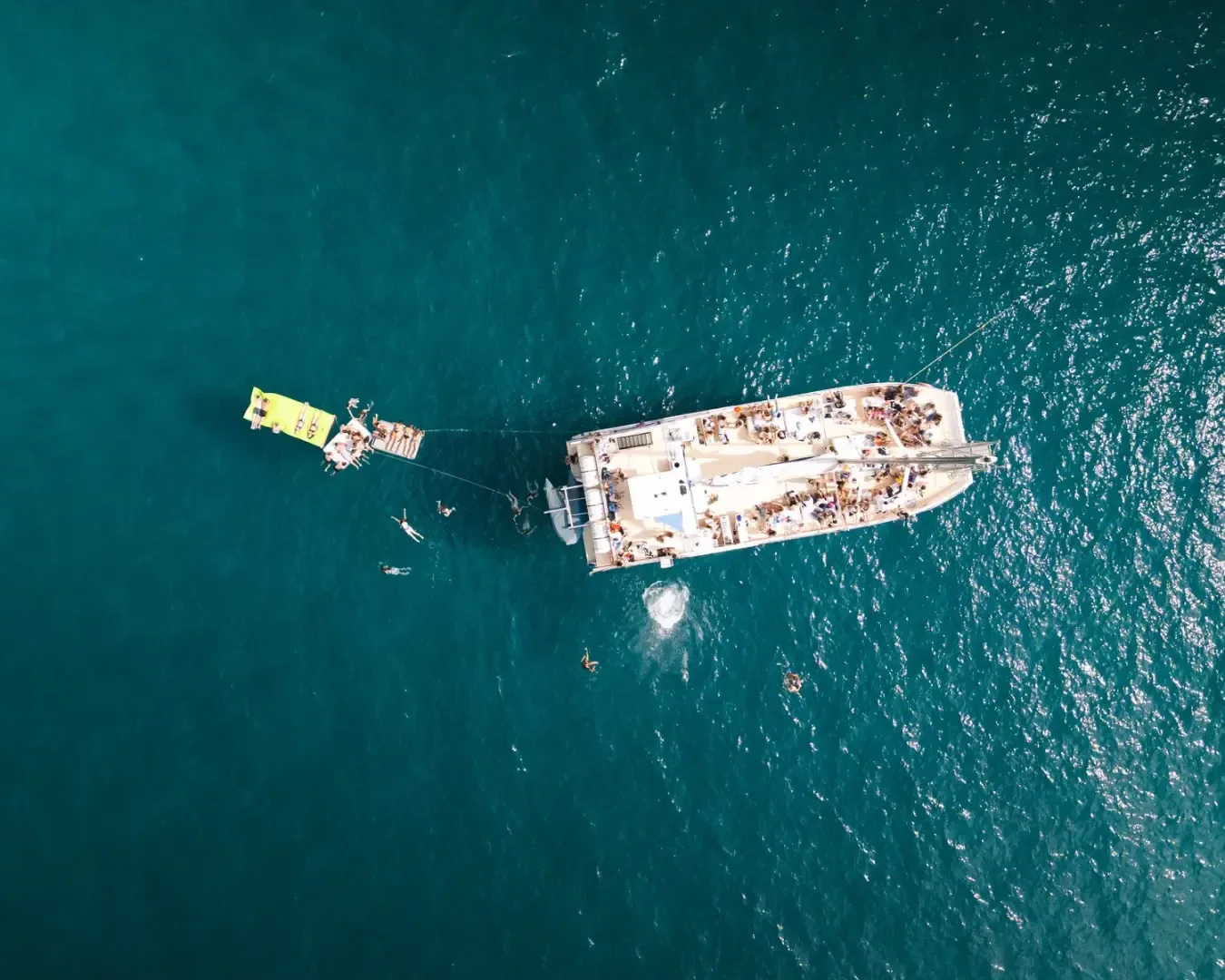 Luftaufnahme eines überfüllten weißen Bootes, das in klarem, blauem Wasser vor Anker liegt, mit schwimmenden Menschen in der Nähe und einer schwimmenden Plattform, die mit dem Boot verbunden ist.
