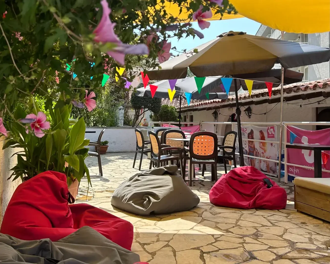 Terrasse im Freien mit Sitzsäcken, Topfpflanzen und Stühlen um Tische unter großen Sonnenschirmen. Bunte Wimpel und rosa Blumen schaffen eine festliche, entspannte Atmosphäre auf einem Steinboden.
