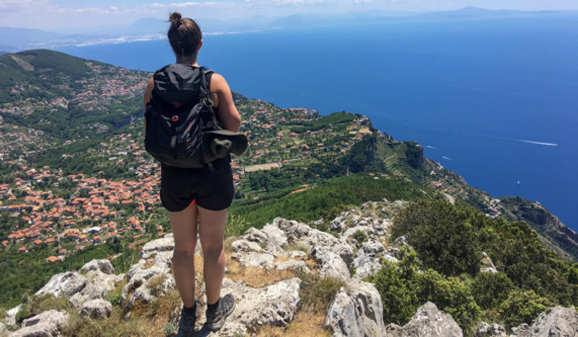 Eine Frau, die einen Rucksack trägt, steht auf einer felsigen Klippe mit Blick auf eine Küstenstadt, grüne Hügel und ein weites blaues Meer unter einem klaren Himmel.