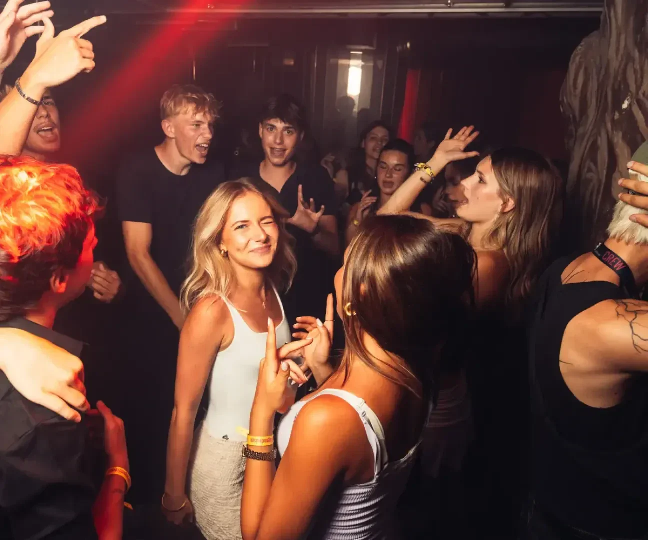 Eine Gruppe junger Erwachsener tanzt und lächelt auf einer überfüllten Indoor-Party, die von warmen, bunten Lichtern beleuchtet wird. Einige Leute heben ihre Arme und genießen die lebhafte Atmosphäre.