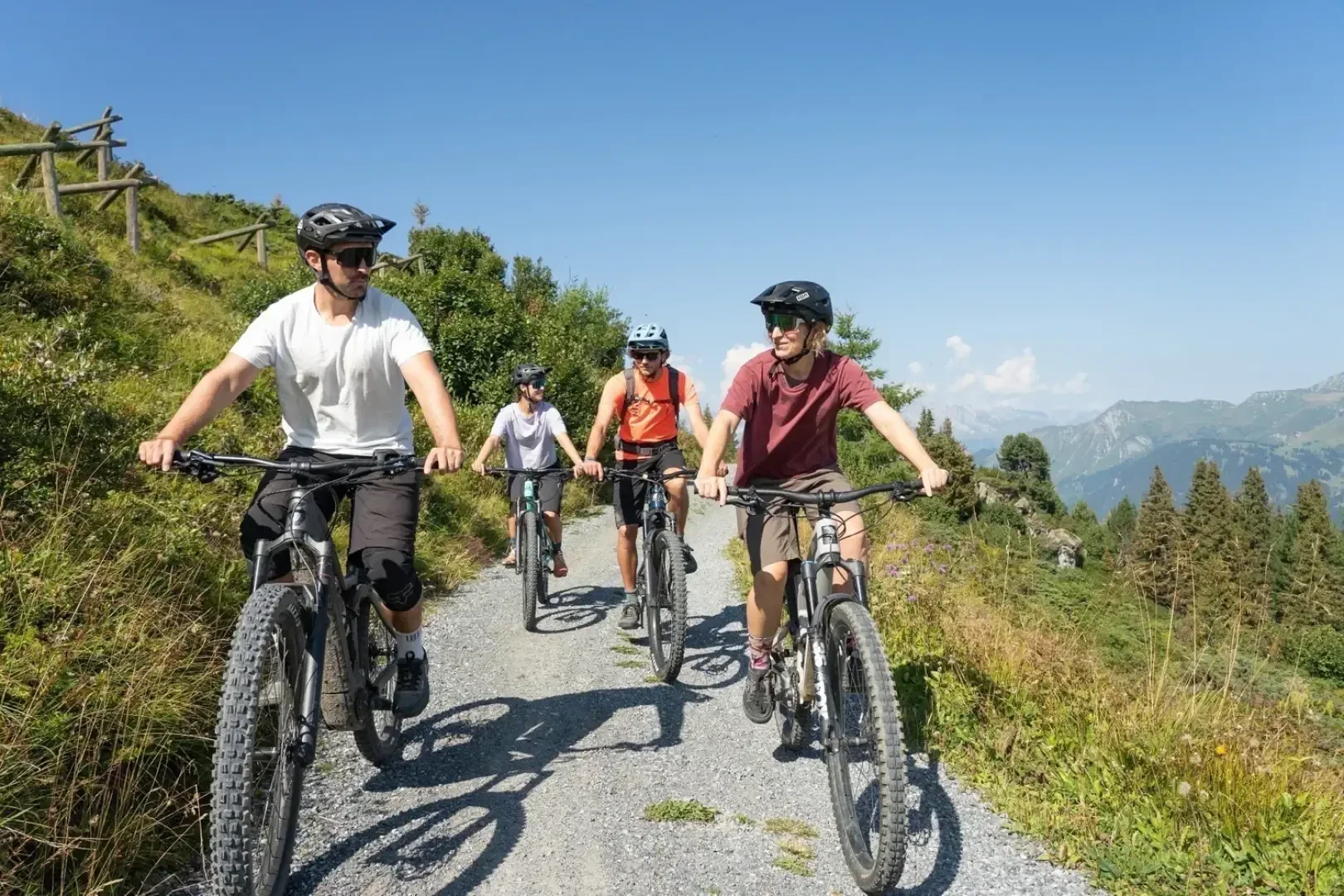 Vier Personen mit Helmen fahren mit dem Mountainbike auf einem Schotterweg durch eine grüne, hügelige Landschaft mit Bäumen und fernen Bergen unter einem klaren blauen Himmel.