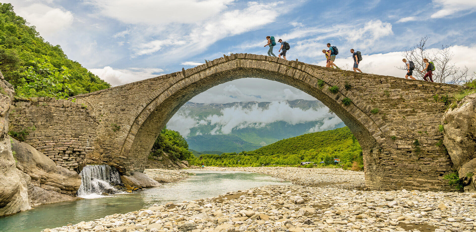 Fünf Wanderer mit Rucksäcken überqueren eine alte Steinbogenbrücke über einen Fluss, umgeben von grünen Hügeln und Bergen mit Wolken im Hintergrund. Die Szene ist hell und natürlich, mit einem Wasserfall neben der Brücke.